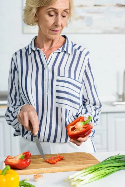 Attractive grey hair woman cutting red bell pepper on wooden board in kitchen — Stock Photo