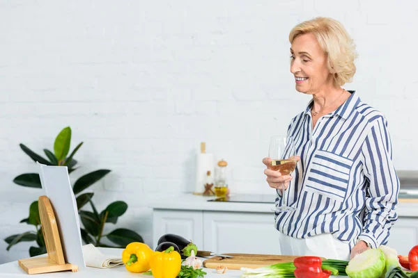 Sorridente attraente donna capelli grigi in possesso di un bicchiere di vino e guardando libro di ricette in cucina — Foto stock