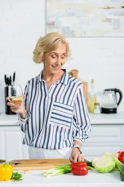 Sonriente atractiva mujer mayor sosteniendo vaso de vino blanco en la cocina y mirando las verduras - foto de stock