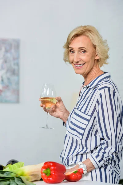 Smiling attractive senior woman holding glass of white wine in kitchen and looking at camera — Stock Photo