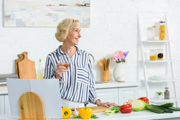 Sonriente atractiva mujer mayor sosteniendo vaso de vino blanco en la cocina y mirando hacia otro lado - foto de stock