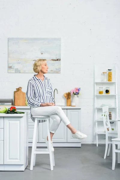 Attractive grey hair woman sitting on high chair in kitchen and looking away — Stock Photo