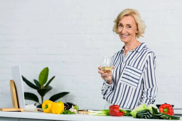 Sonriente atractiva mujer mayor sosteniendo vaso de vino blanco en la cocina - foto de stock