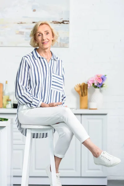 Atractiva mujer de pelo gris sentado en la silla alta en la cocina y mirando a la cámara - foto de stock