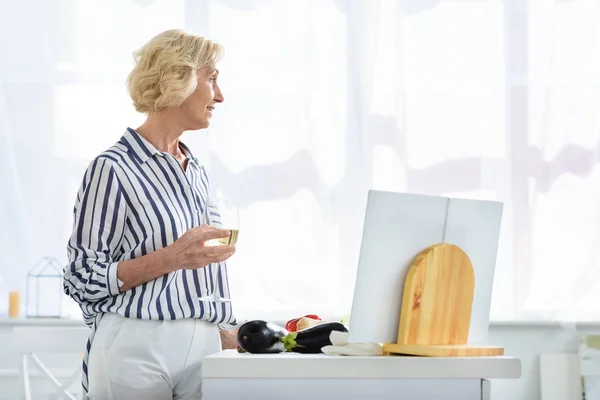 Vista lateral de la atractiva mujer de pelo gris sosteniendo un vaso de vino en la cocina y mirando hacia otro lado - foto de stock