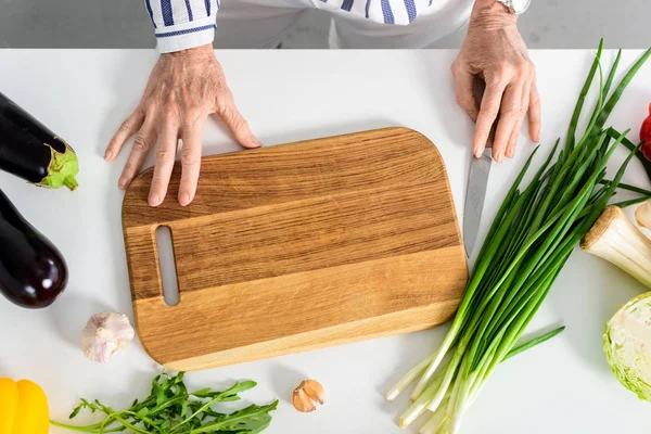 Cropped image of senior woman touching wooden board and knife in kitchen — Stock Photo