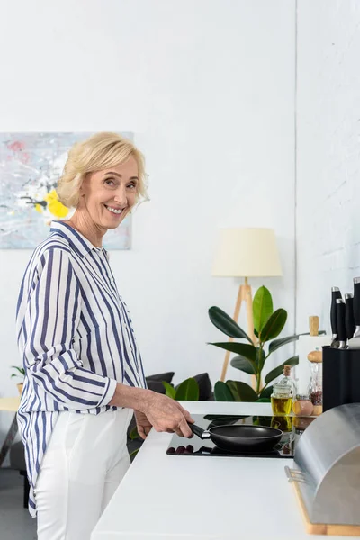Smiling attractive grey hair woman cooking in kitchen and putting frying pan on stove — Stock Photo