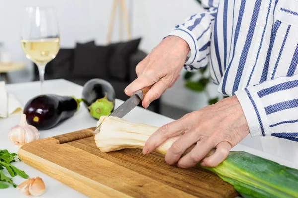 Cropped image of grey hair woman cutting leek on wooden board in kitchen — Stock Photo