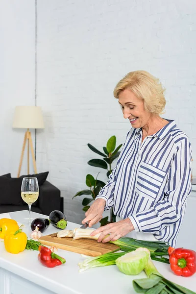 Smiling attractive grey hair woman cutting leek on wooden board in kitchen — Stock Photo