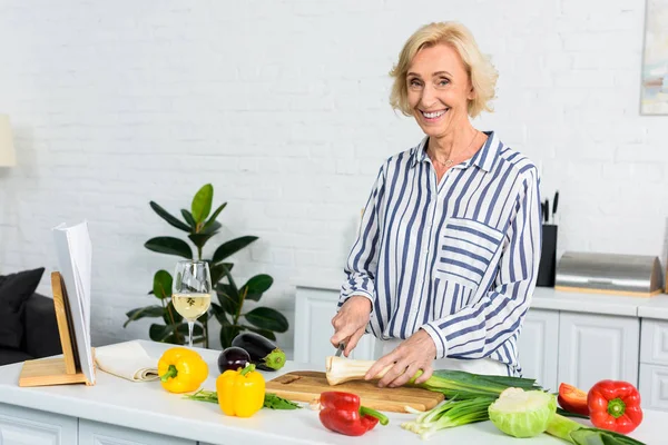 Lächelnde grauhaarige Frau schneidet Lauch auf Holzbrett in der Küche und blickt in die Kamera — Stockfoto