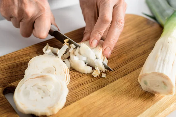 Cropped image of senior woman cutting ripe leek on wooden board in kitchen — Stock Photo