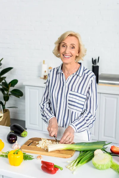 Souriant attrayant senior femme coupe poireau sur planche en bois dans la cuisine et en regardant la caméra — Photo de stock