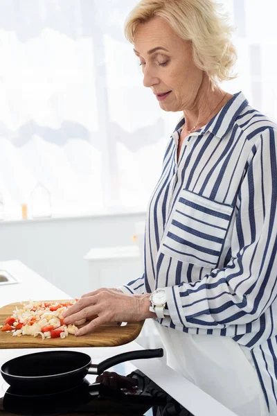 Side view of attractive senior woman putting vegetables on frying pan in kitchen — Stock Photo