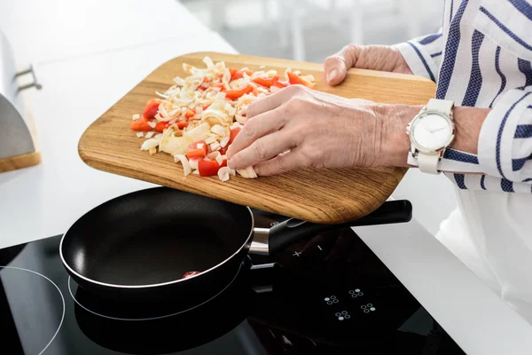 Imagen recortada de mujer mayor poniendo verduras en la sartén en la cocina - foto de stock