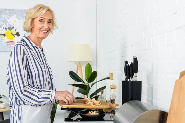 Side view of smiling attractive senior woman putting vegetables on frying pan in kitchen — Stock Photo