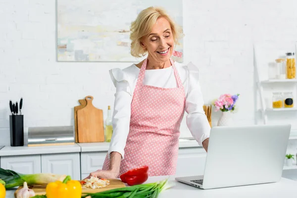 Sonriente atractiva mujer mayor cocina y el uso de ordenador portátil en la cocina - foto de stock