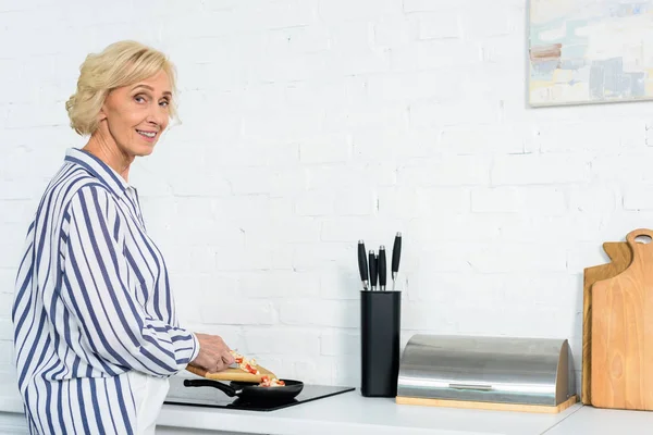 Vista lateral de la sonriente mujer de pelo gris poniendo verduras en la sartén en la cocina y mirando a la cámara - foto de stock