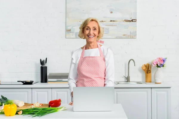 Усміхнена приваблива старша жінка використовує ноутбук для приготування їжі на кухні і дивиться на камеру — стокове фото