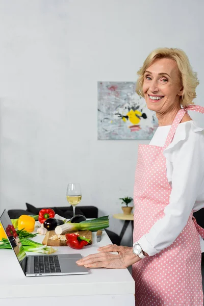Side view of smiling attractive senior woman using laptop for cooking in kitchen — Stock Photo