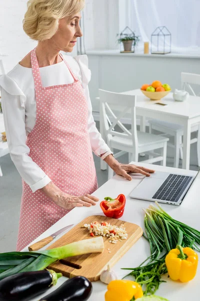 Belle femme âgée utilisant un ordinateur portable pour cuisiner dans la cuisine — Photo de stock