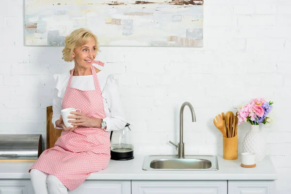 Attractive senior woman sitting on kitchen counter with cup of coffee and looking away — Stock Photo