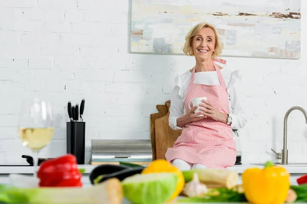 Lächelnde schöne Seniorin, die mit einer Tasse Tee auf der Küchentrese sitzt und in die Kamera blickt — Stockfoto