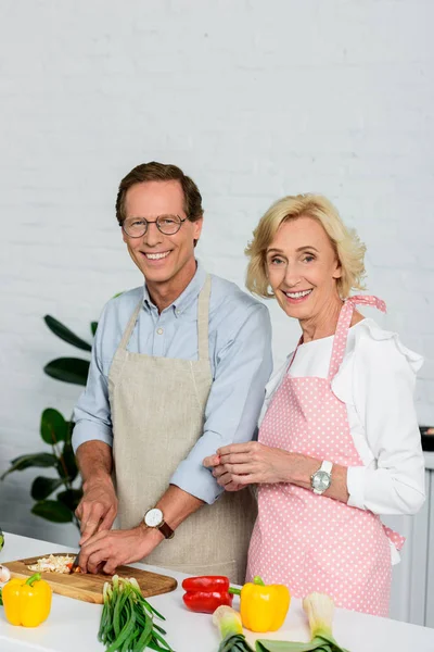 Усміхнена старша пара готує разом на кухні і дивиться на камеру — стокове фото