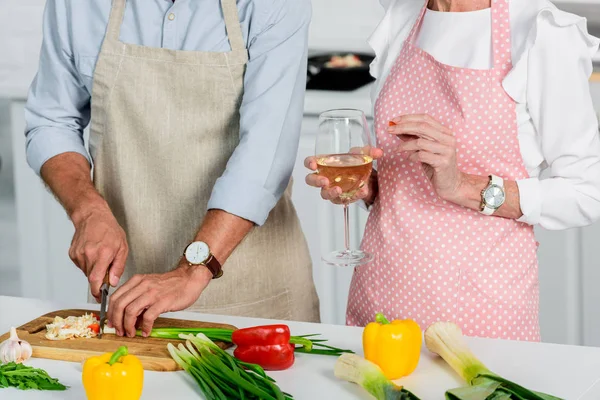 Imagen recortada de marido mayor cortando verduras y esposa sosteniendo un vaso de vino en la cocina - foto de stock
