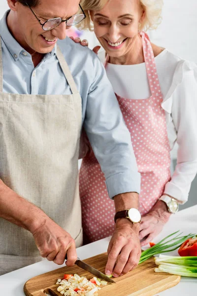 Sonriendo esposa mayor abrazando marido mientras él corte cebolla verde en la cocina - foto de stock