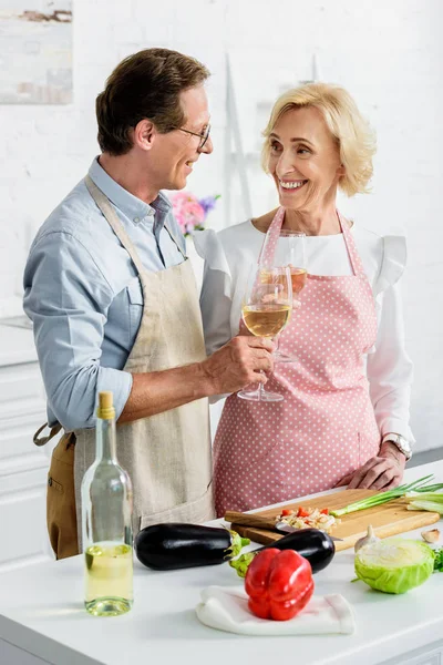 Улыбающаяся пожилая пара, звенящая с бокалами вина во время приготовления пищи на кухне и глядя друг на друга — стоковое фото
