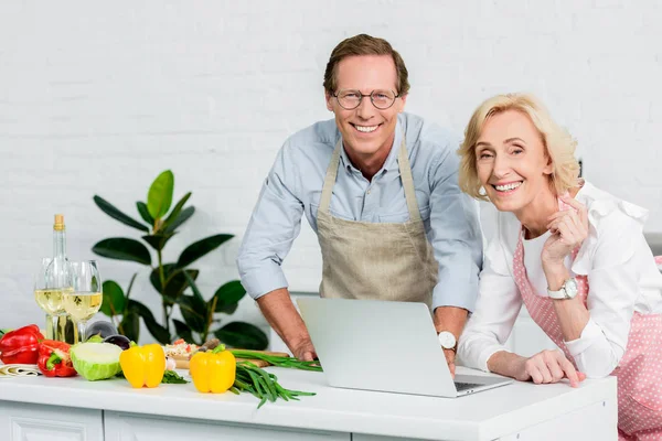 Pareja de ancianos cocinar y el uso de ordenador portátil para recetas en la cocina - foto de stock