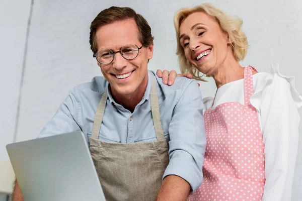 Low angle view of smiling senior couple in aprons looking at laptop at kitchen — Stock Photo