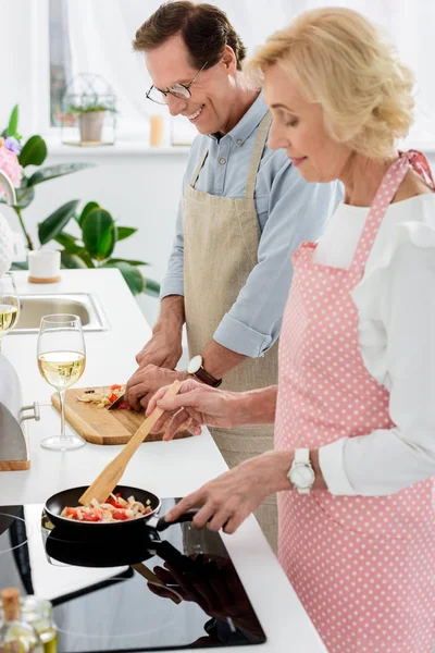 Side view of senior couple cooking vegetables on frying pan and cutting vegetables on wooden board at kitchen — Stock Photo