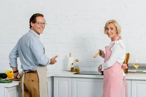 Sorrindo esposa sênior e marido cozinhar juntos na cozinha e olhando para a câmera — Fotografia de Stock