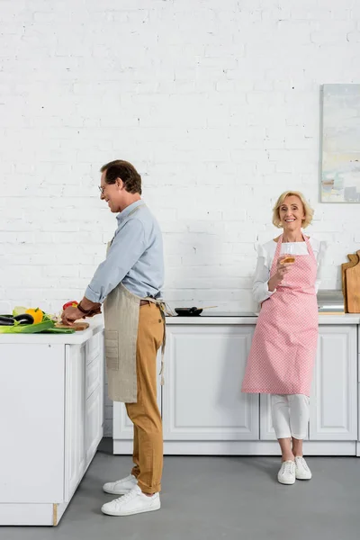 Marito anziano cucina in cucina e moglie in piedi con un bicchiere di vino — Foto stock