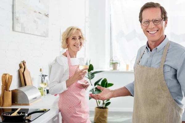 Feliz anciano en delantal y anteojos sonriendo a la cámara mientras su esposa bebe vino en la cocina - foto de stock