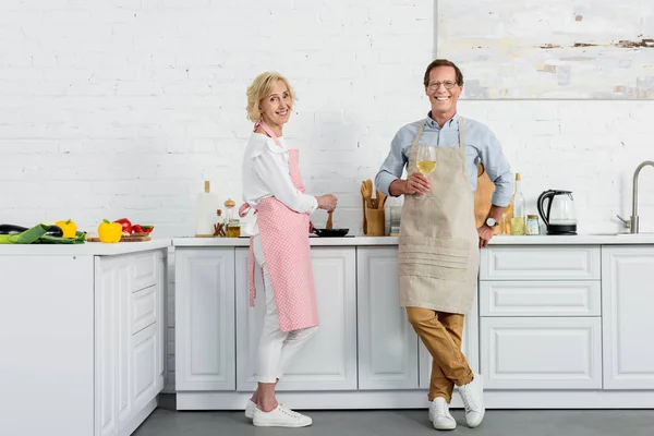 Felice coppia anziana in grembiuli sorridente alla macchina fotografica mentre beve vino e cucina in cucina — Foto stock