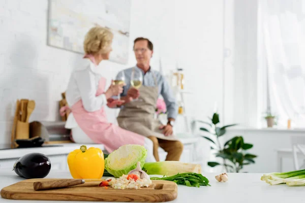 Close-up view of fresh vegetables in wooden cutting board and senior couple drinking wine behind in kitchen — Stock Photo