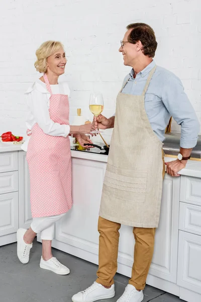 Feliz pareja de ancianos bebiendo vino y sonriéndose mientras cocinan juntos en la cocina - foto de stock