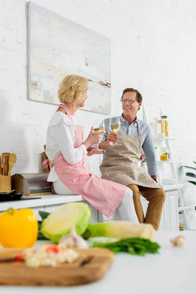Happy elderly couple in aprons holding glasses of wine and smiling each other in kitchen — Stock Photo