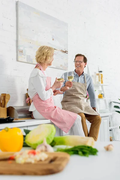 Feliz casal de idosos em aventais segurando copos de vinho e sorrindo uns aos outros na cozinha — Fotografia de Stock