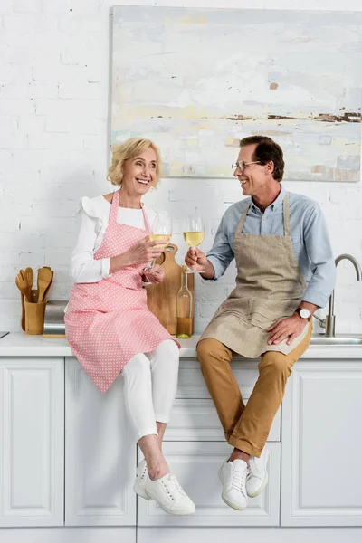 Щаслива старша пара в фартухах тримає келихи вина, сидячи разом на кухні — стокове фото