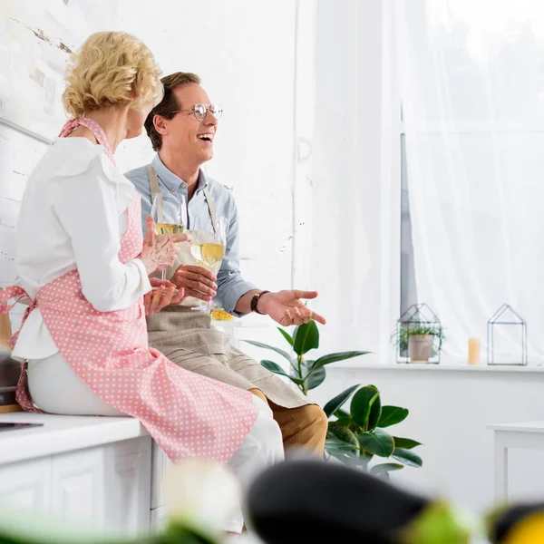 Glückliches Seniorenpaar in Schürzen trinkt Wein und lacht gemeinsam in der Küche — Stockfoto