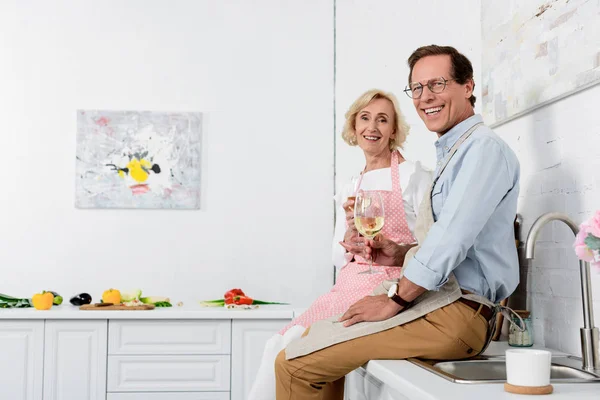 Feliz pareja de ancianos en delantales sosteniendo vasos de vino y sonriendo a la cámara en la cocina - foto de stock