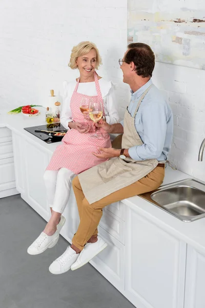 Vista de ángulo alto de feliz pareja de ancianos n delantales beber vino y sonreír unos a otros en la cocina - foto de stock