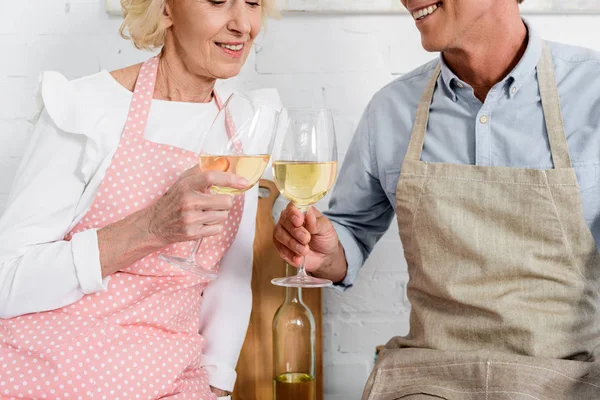 Обрезанный снимок счастливой пожилой пары в фартуках, пьющей вино во время приготовления пищи на кухне — стоковое фото
