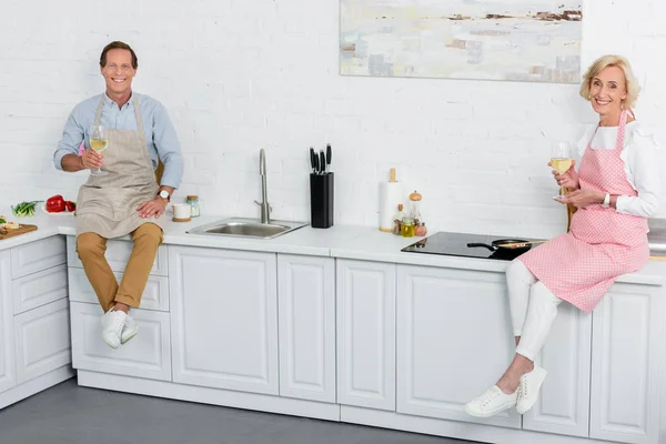 Heureux couple âgé dans des tabliers tenant des verres de vin et souriant à la caméra dans la cuisine — Photo de stock