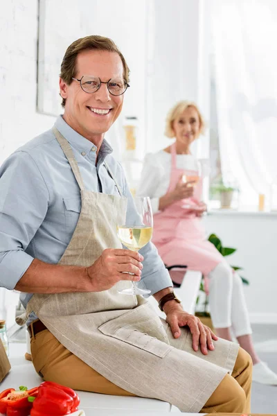 Happy senior couple holding glasses of wine and smiling at camera while cooking together — Stock Photo
