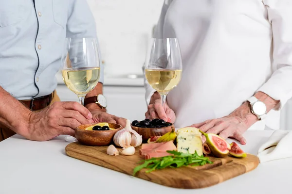 Primer plano vista parcial de pareja de ancianos sosteniendo vasos de vino mientras disfruta de deliciosos aperitivos en casa - foto de stock