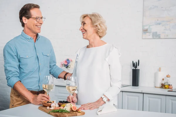 Belo casal feliz idosos conversando e sorrindo uns aos outros enquanto bebe vinho e comer deliciosos lanches em casa — Fotografia de Stock